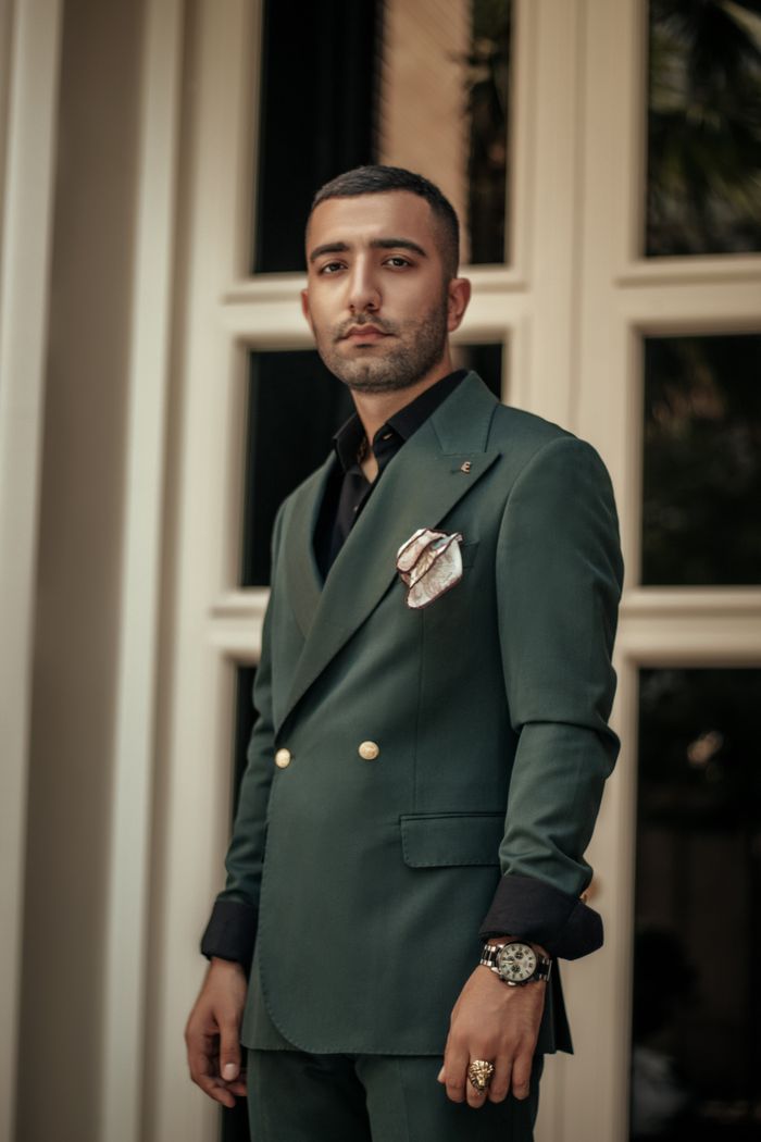 Elegant portrait of a man in a green suit and black shirt, standing against a window.
