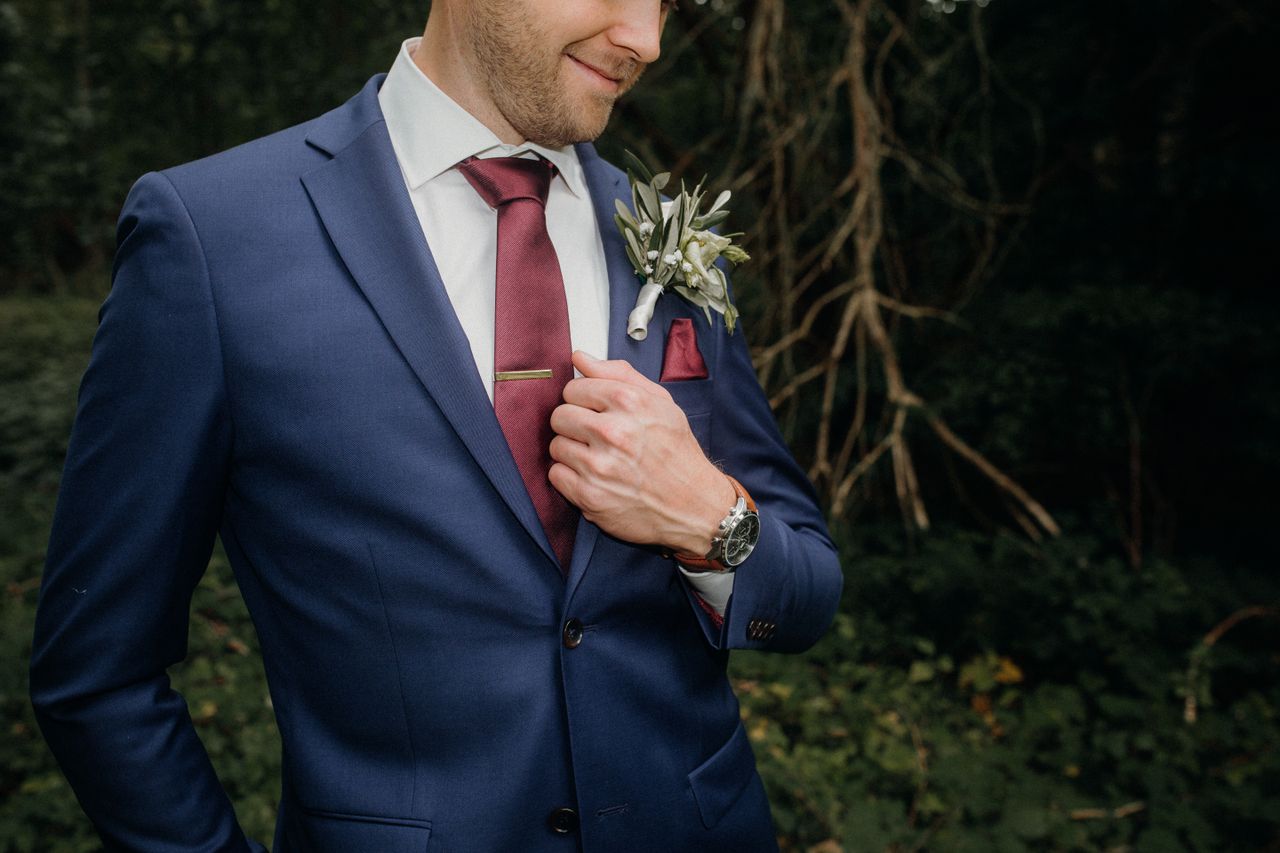 Man in navy suit with red tie and boutonniere standing outdoors, exuding elegance and style.