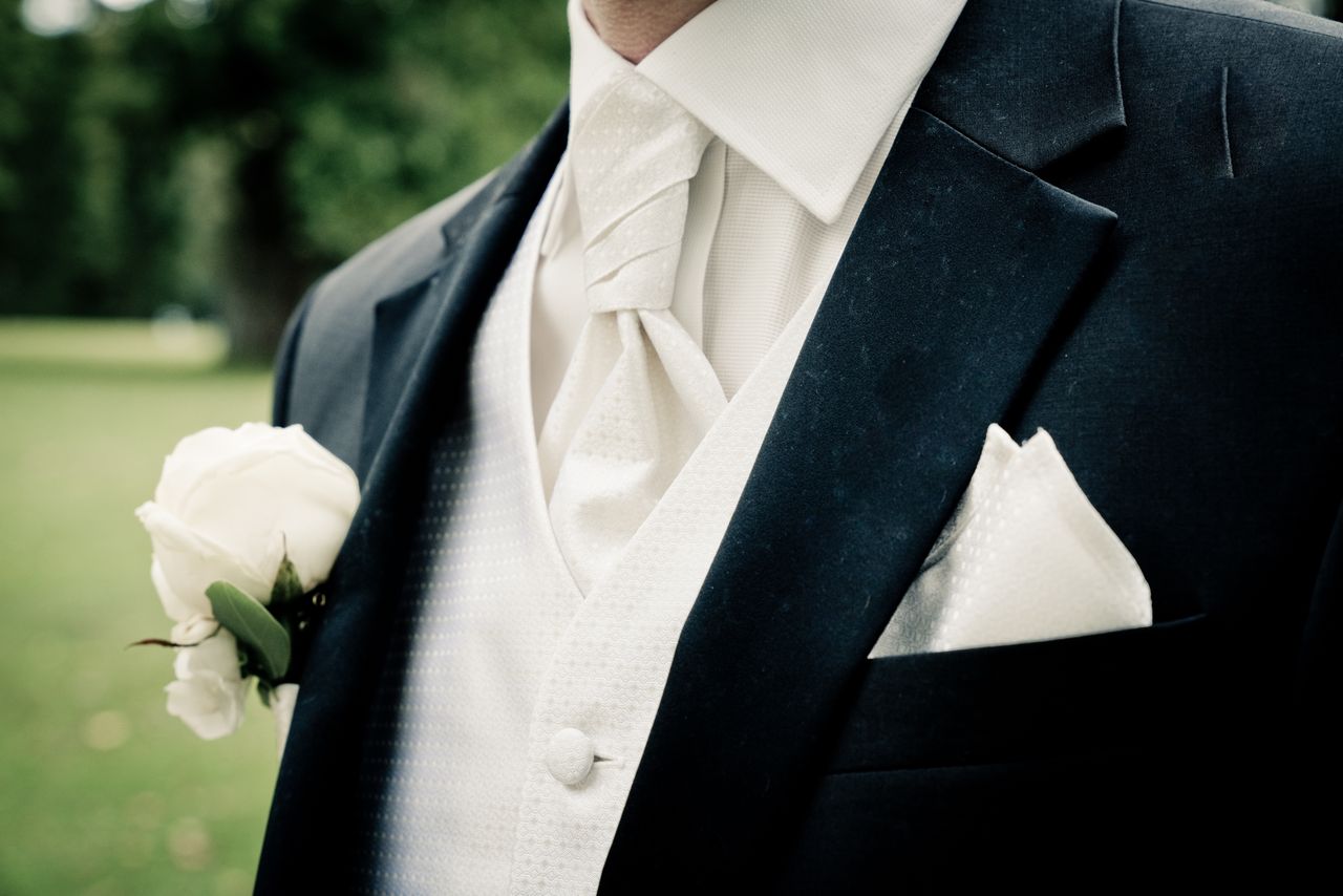 about-us Close-up of groom's suit and tie with white rose boutonniere, symbolizing wedding elegance.