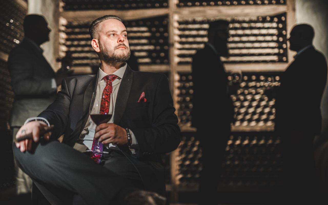 Elegant man in a suit enjoying a drink in a wine cellar, surrounded by shadows.