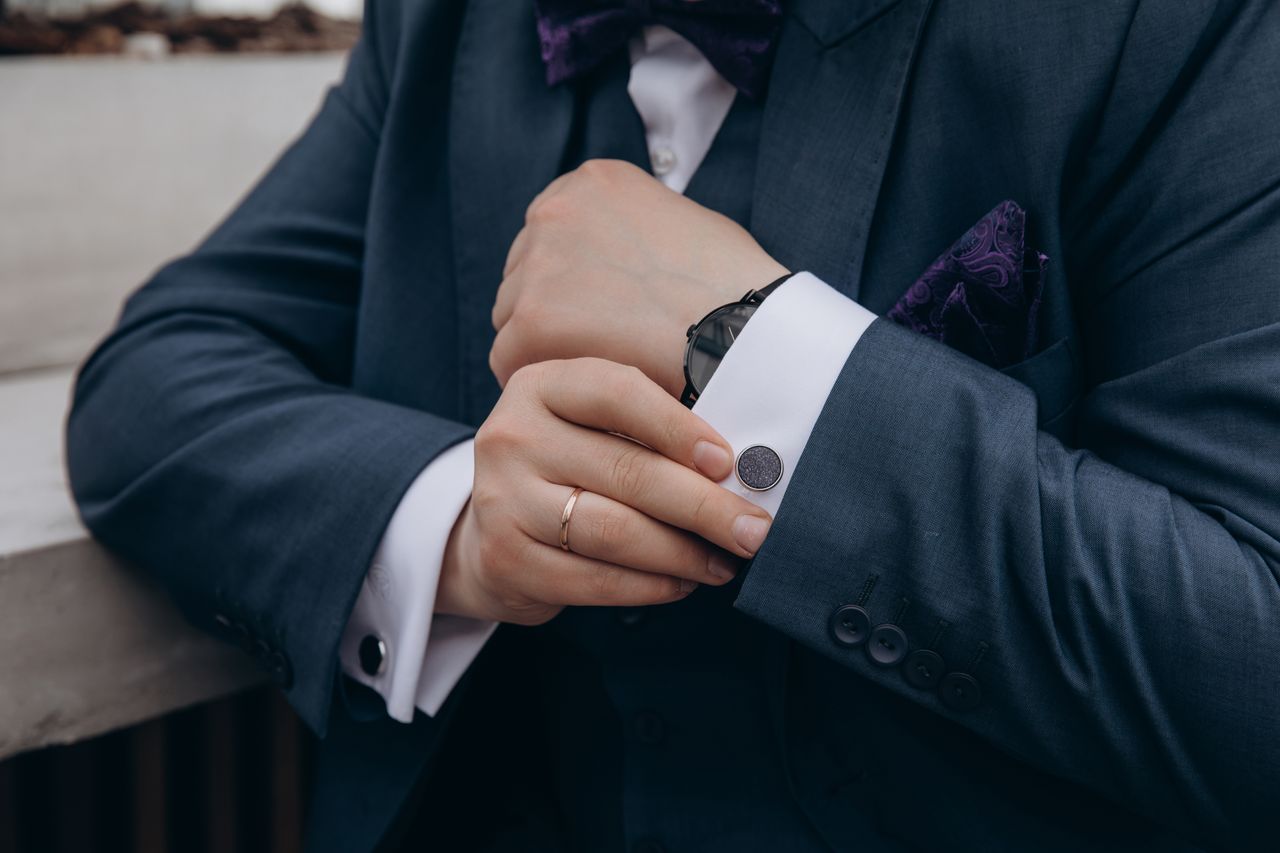 our-story Close-up of a man adjusting his suit jacket cuff with a watch and ring visible, showing elegance.